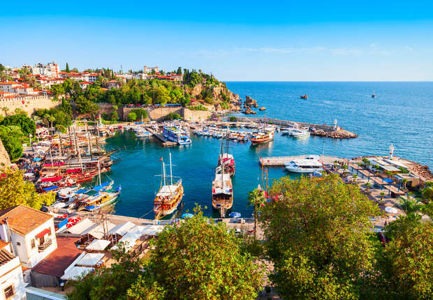 Boats are docked in a colorful, bustling harbor surrounded by lush greenery and historical architecture. The scene is set against a bright blue ocean, with clear skies above, creating a vibrant coastal view.